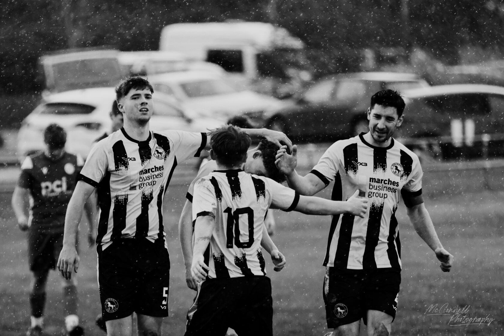 Football players in black and white striped jerseys celebrating together in the rain, with one player having their arm raised in celebration. The scene captures intensity and camaraderie amidst a downpour.