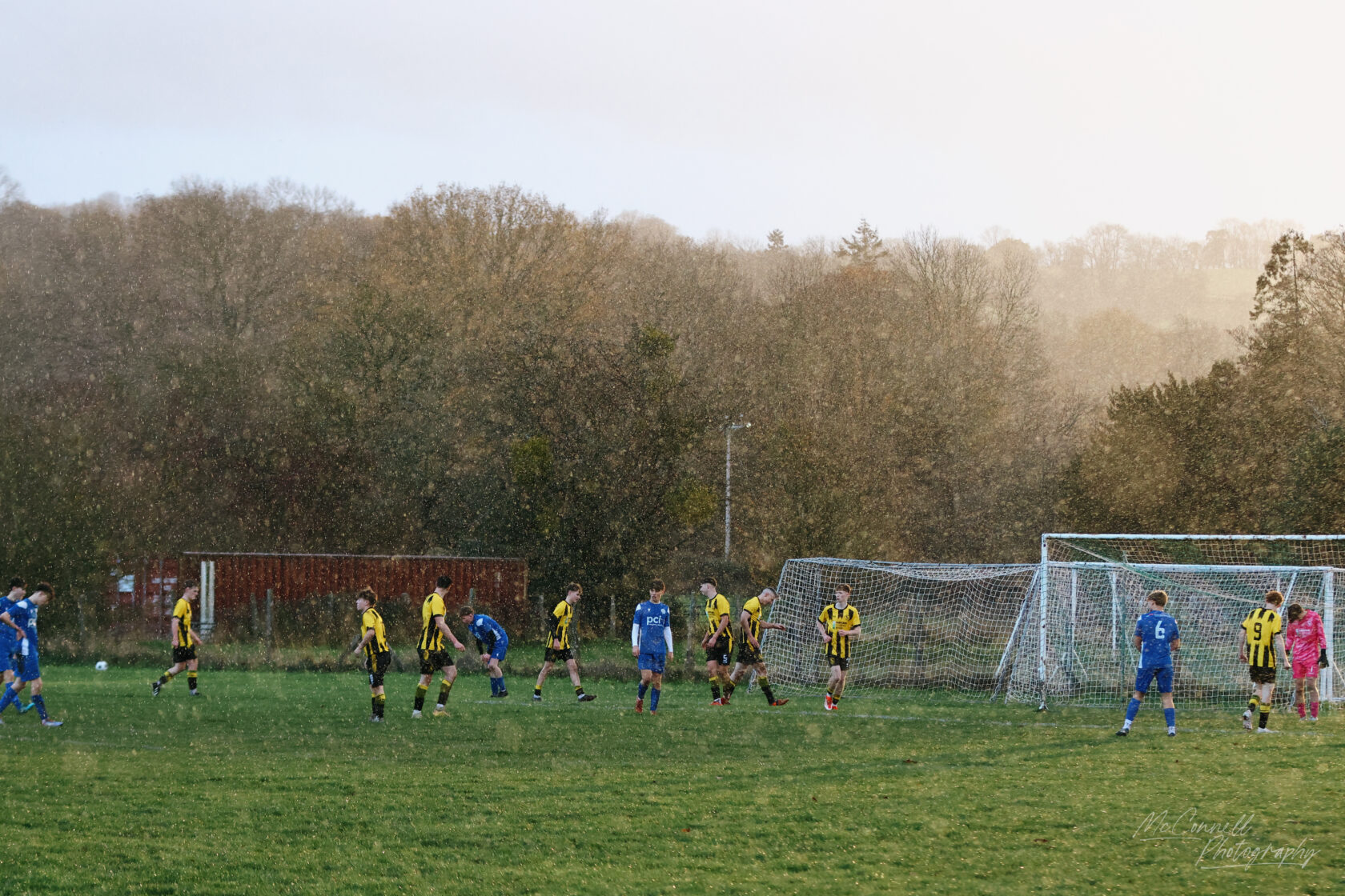 A youth football match taking place on a grassy field under heavy rain, with players in yellow and black striped jerseys opposing those in blue. The goal posts are visible in the background.