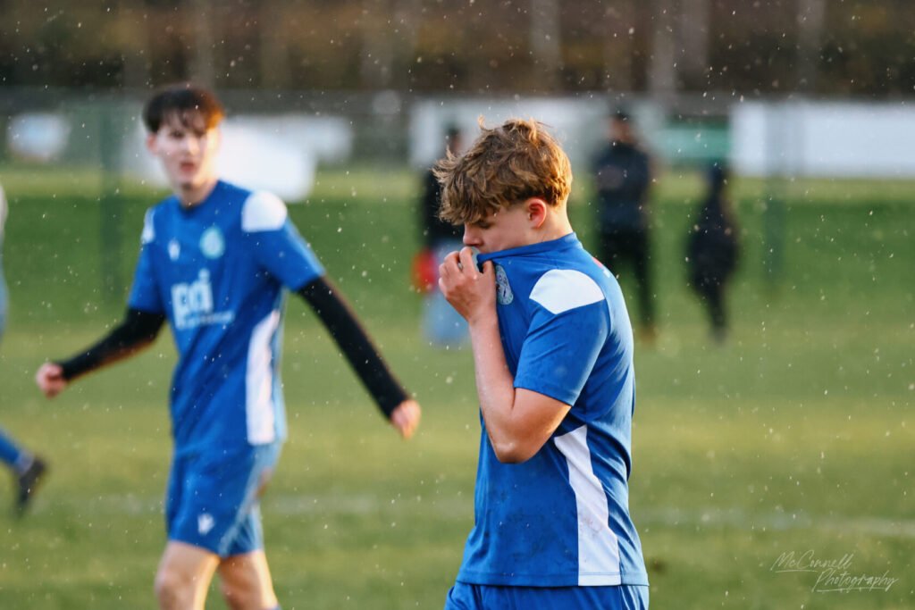 A young football player in a blue jersey stands on a football pitch, looking dejected as he pulls his shirt up to his face, while another player in the background walks away.