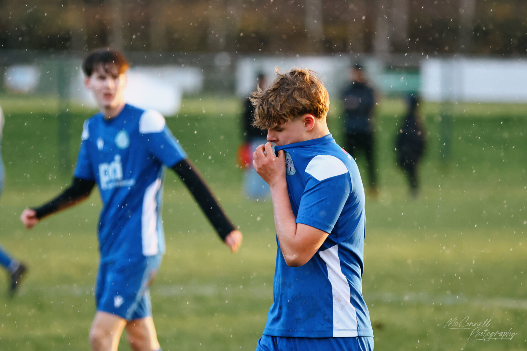 A young football player in a blue jersey stands on a football pitch, looking dejected as he pulls his shirt up to his face, while another player in the background walks away.