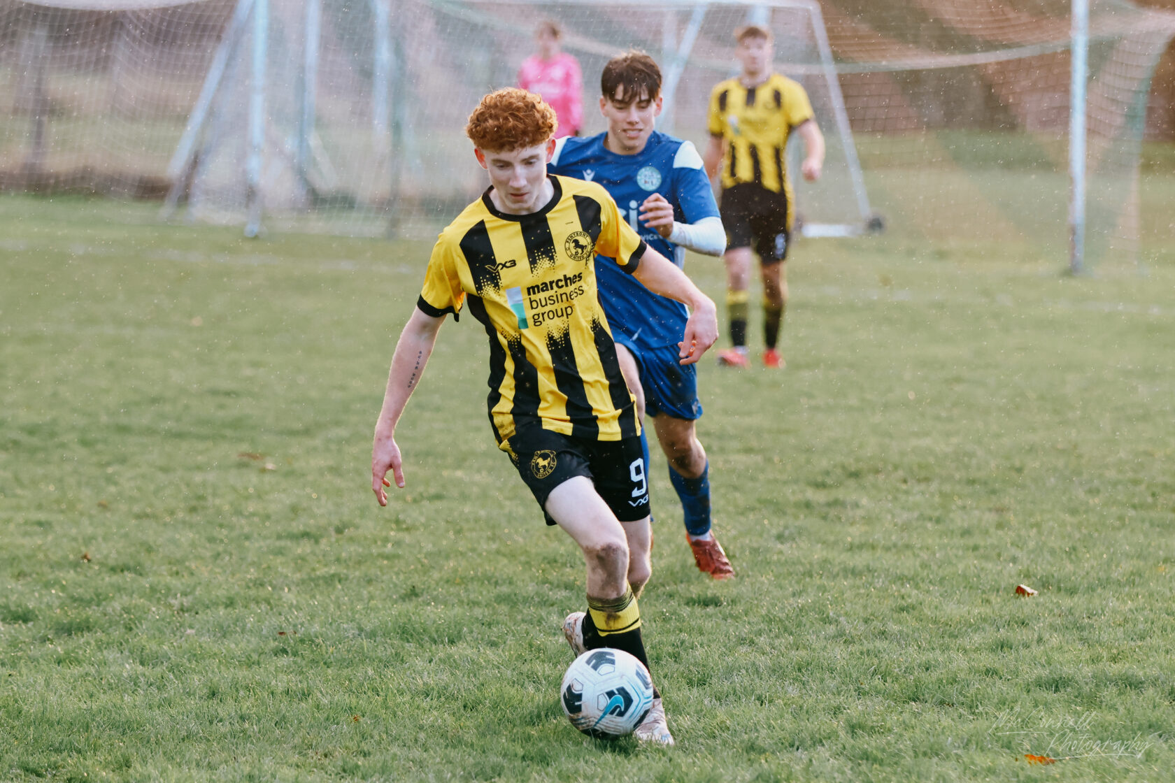 A young football player in a black and yellow striped kit dribbles a ball on a grassy pitch, with another player in blue chasing behind. The scene captures the energy of a youth football match.
