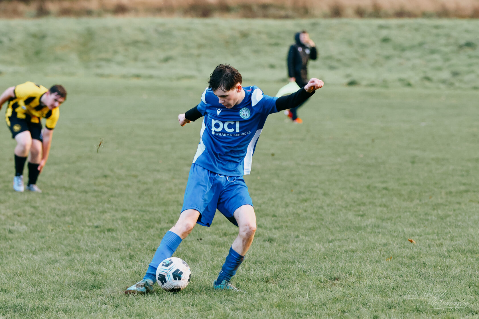 A young male football player in a blue kit is striking a soccer ball on a grassy field, with another player in a yellow and black striped kit in the background.
