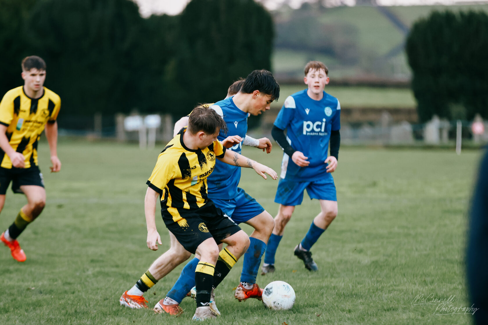 Two teams compete in a youth football match, with players in yellow and black stripes battling against those in blue. The focus is on two players closely interacting over the ball in a grassy field.