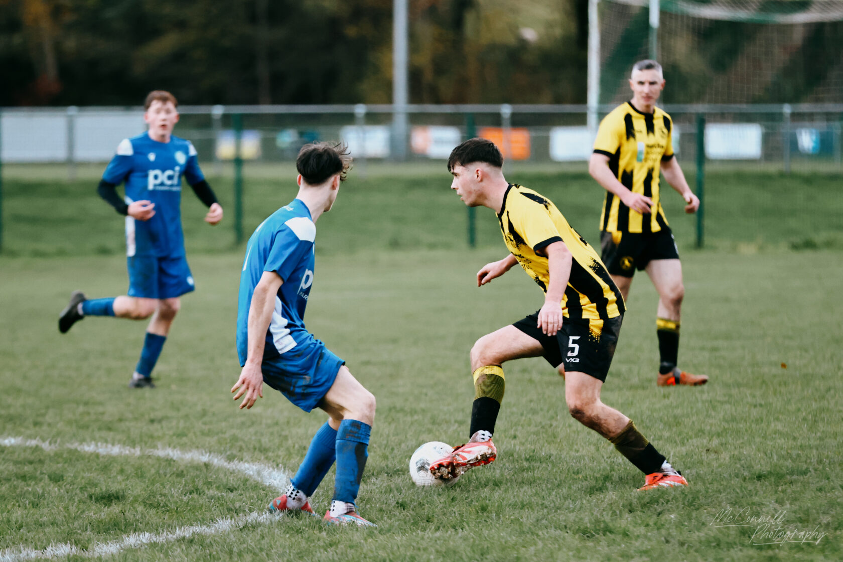 A competitive moment during a football match, with players in blue and yellow kits battling for possession of the ball on the pitch.
