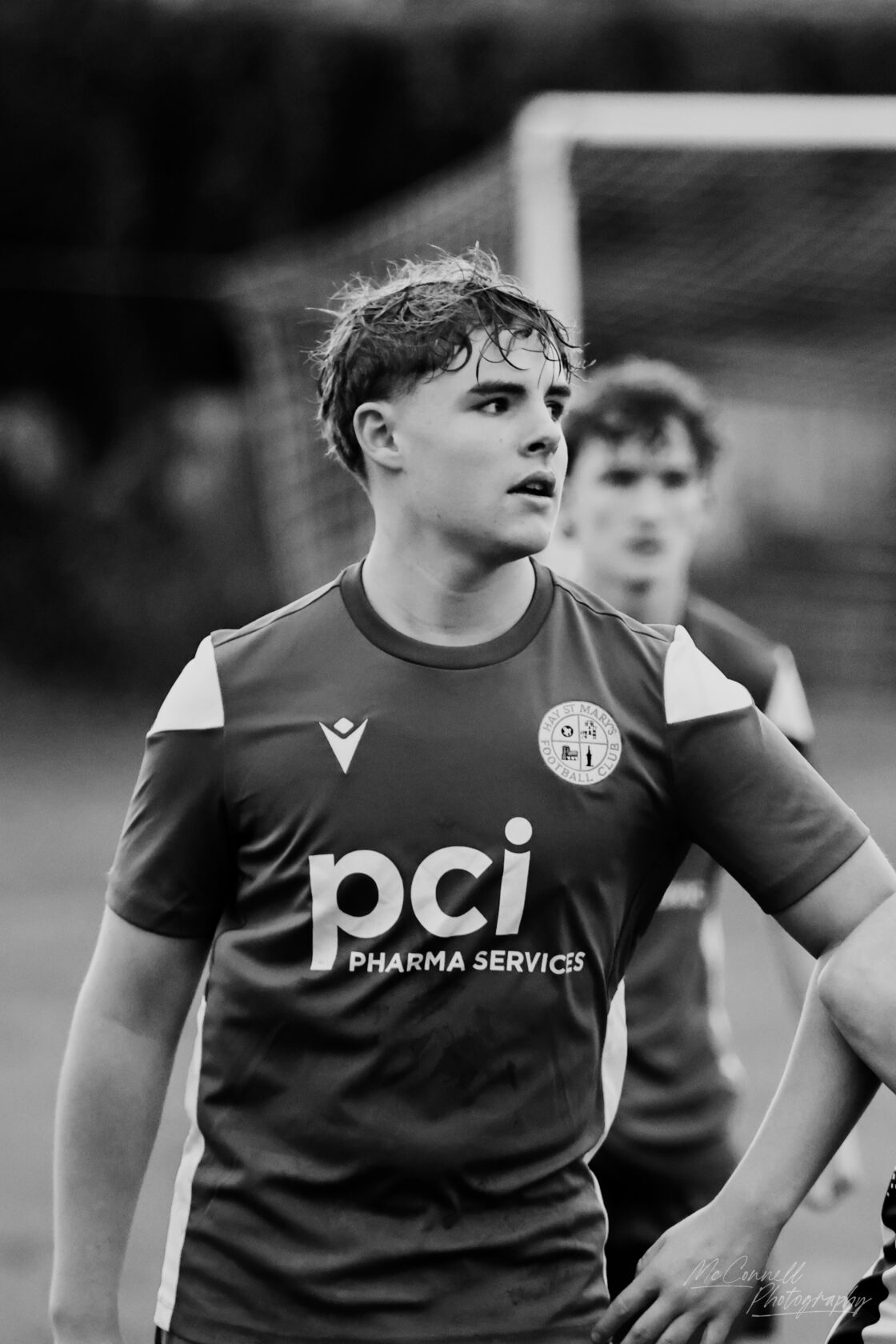 A young male football player wearing a red team jersey with the logo of a football club. He has wet hair and an intense expression, with another player blurred in the background. The photo is in black and white.