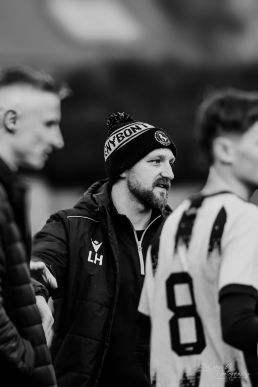 A football coach wearing a beanie with 'NEW BORN' printed on it, interacting with players on the sidelines during a match. The image is in black and white.