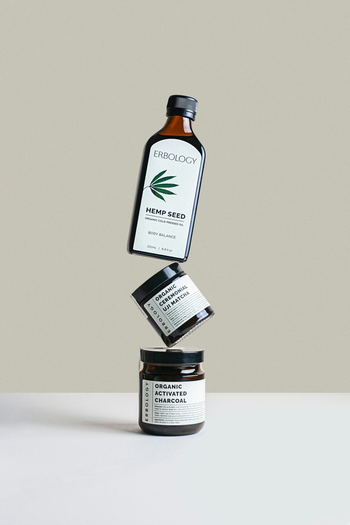 Three Erbology health products arranged in a floating stack, featuring a bottle of organic hemp seed oil at the top, a jar of organic ceremonial uji matcha in the middle, and a jar of organic activated charcoal at the bottom. The background is a light beige colour.