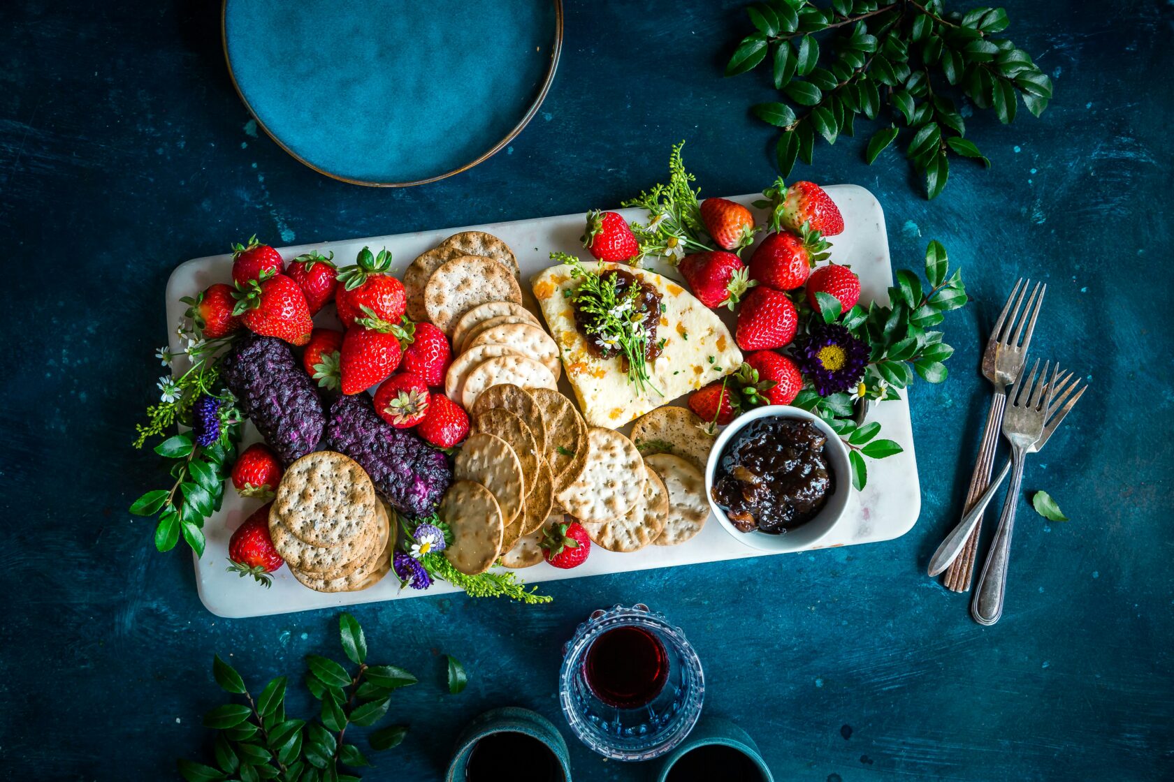 A beautifully arranged platter featuring slices of cheese, assorted crackers, fresh strawberries, a small bowl of dark fruit jam, and decorative herbs, all on a blue textured background.