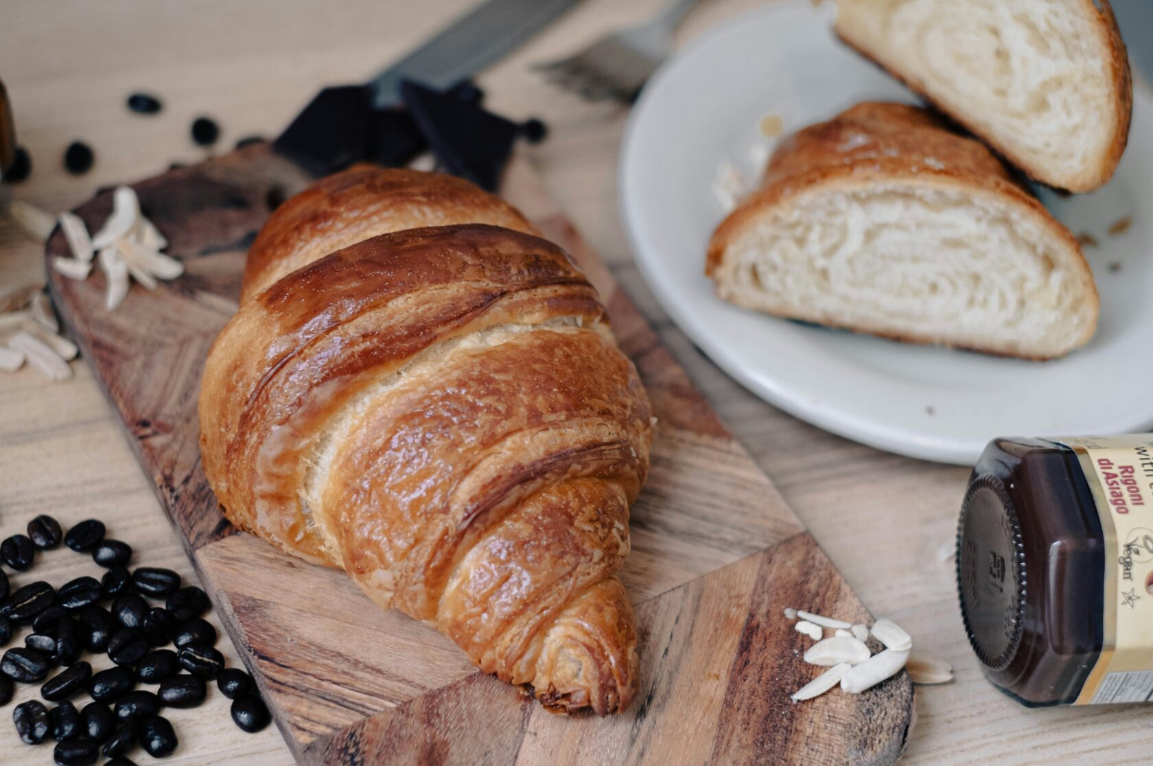 A golden brown croissant resting on a wooden board, with scattered coffee beans, almond slices, and a plate featuring a halved croissant and a jar of chocolate spread in the background.