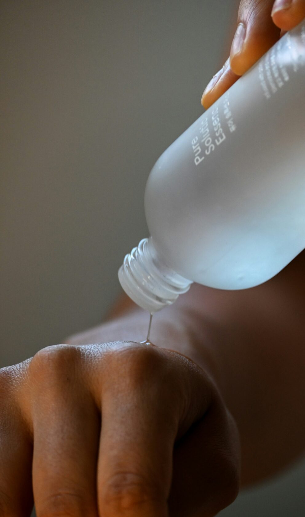 A hand pouring a clear essence from a glass bottle onto the skin of another hand.