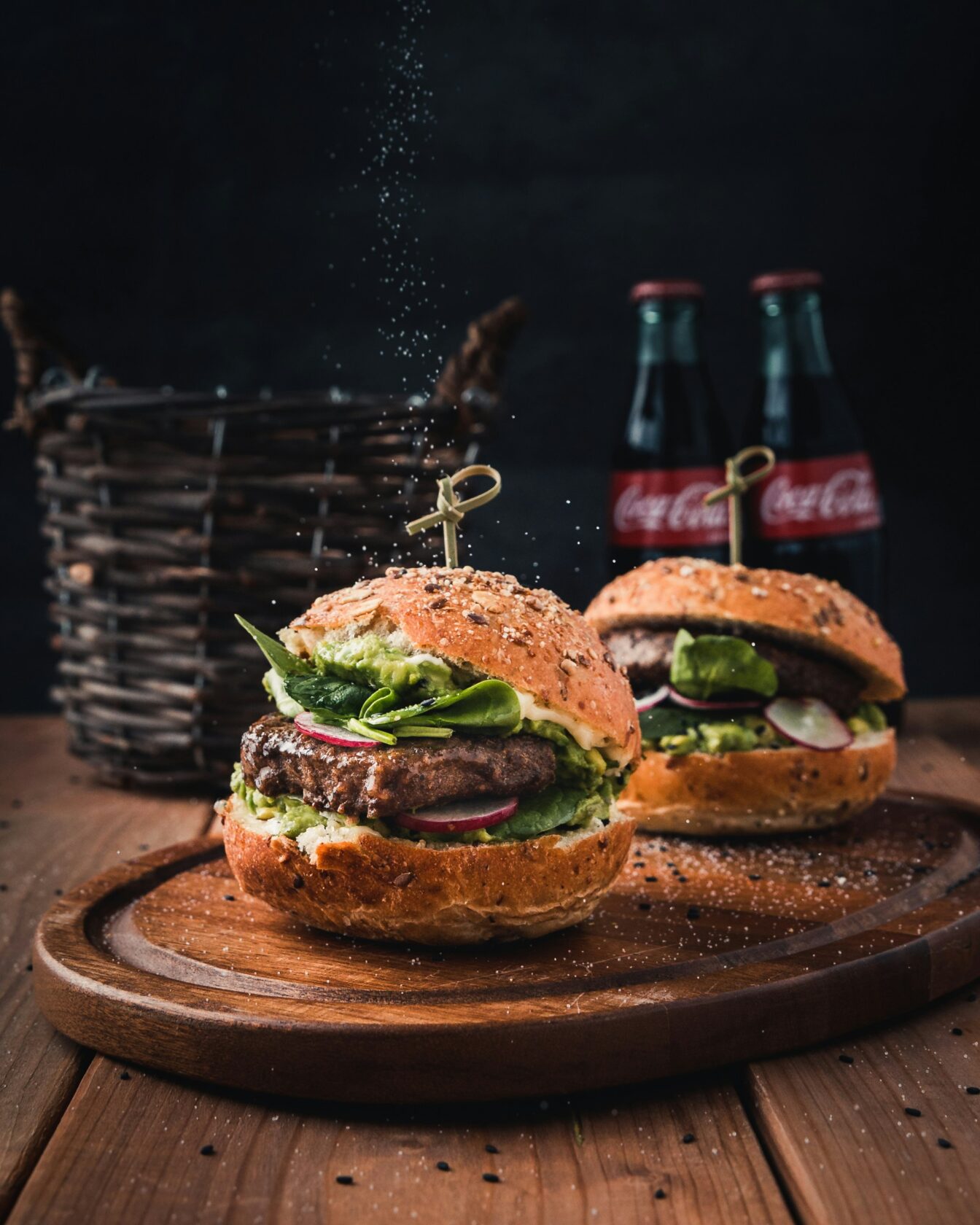 Two gourmet burgers featuring fresh ingredients like lettuce, radish, and avocado on a wooden platter, with bottles of Coca-Cola in the background and seasoning being sprinkled.