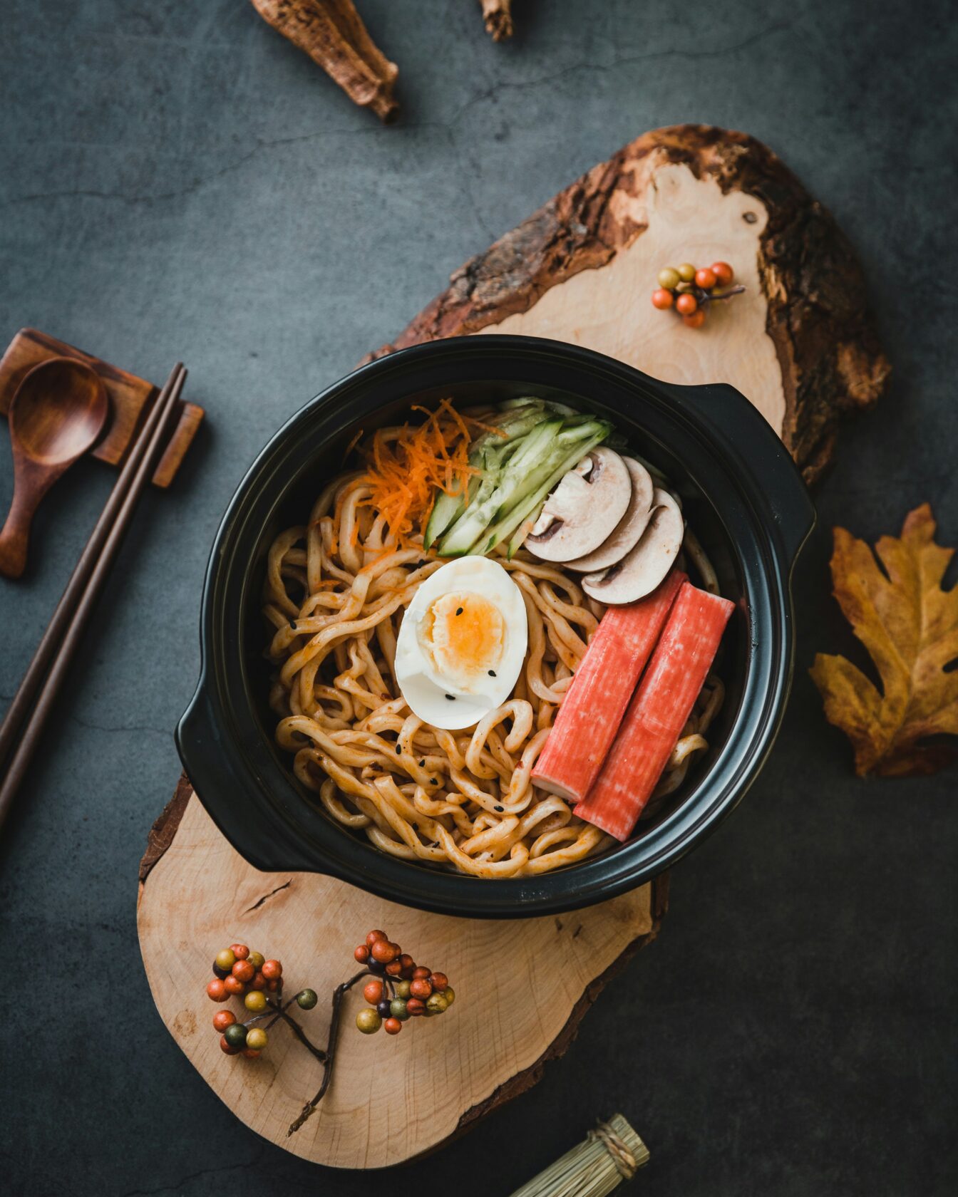 A dark ceramic bowl filled with noodles, topped with a half-boiled egg, crab sticks, cucumber, mushrooms, and shredded carrots, placed on a wooden surface alongside chopsticks and decorative foliage.