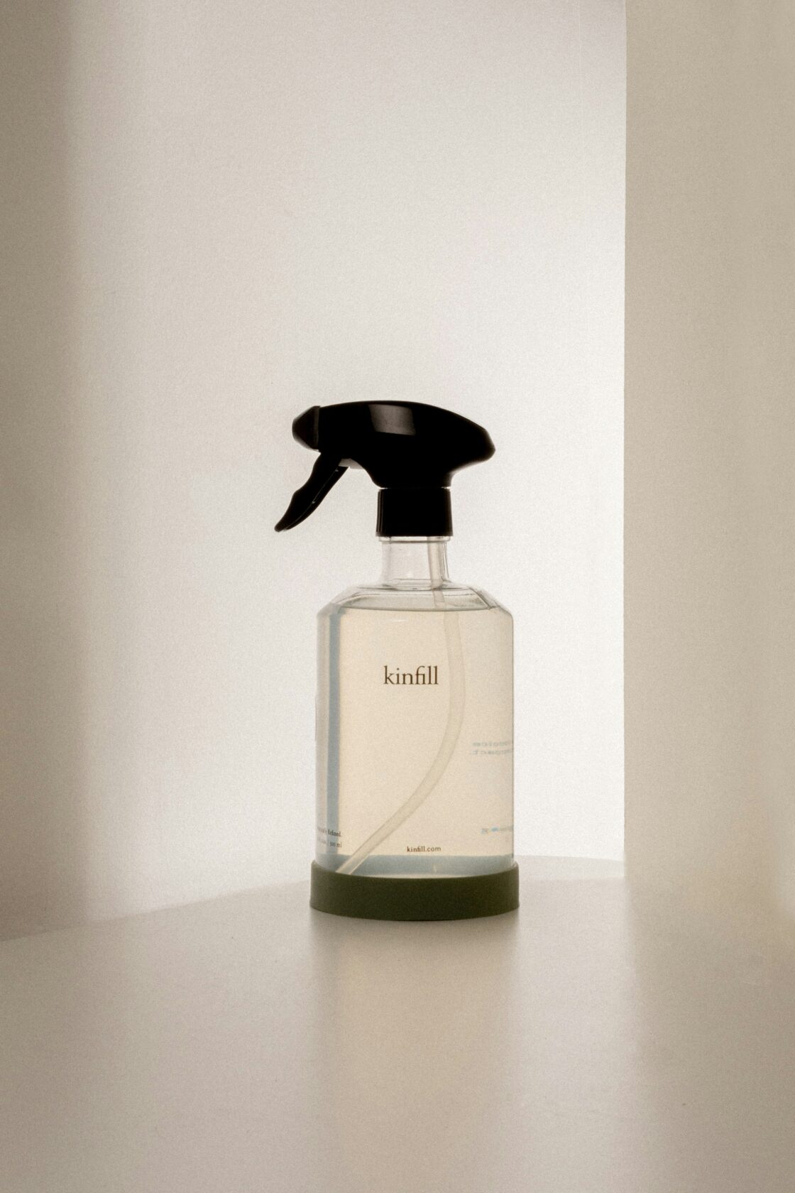 A transparent spray bottle with a black nozzle, featuring a green base, containing a clear liquid, placed on a minimalist white surface.