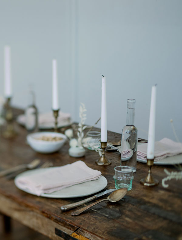 Elegant Dining Table Setup A rustic wooden dining table beautifully arranged with white candles in brass holders, plates with pale pink napkins, glassware, and a small bowl of snacks.