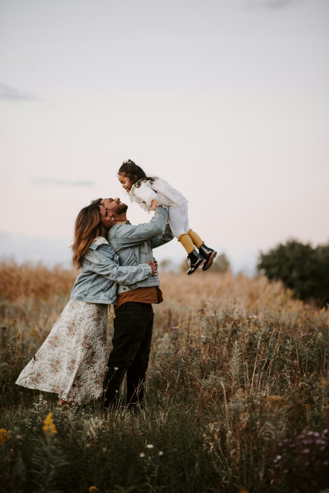 A happy family in a field, with a father lifting his daughter while the mother hugs them, all smiling against a sunset background.