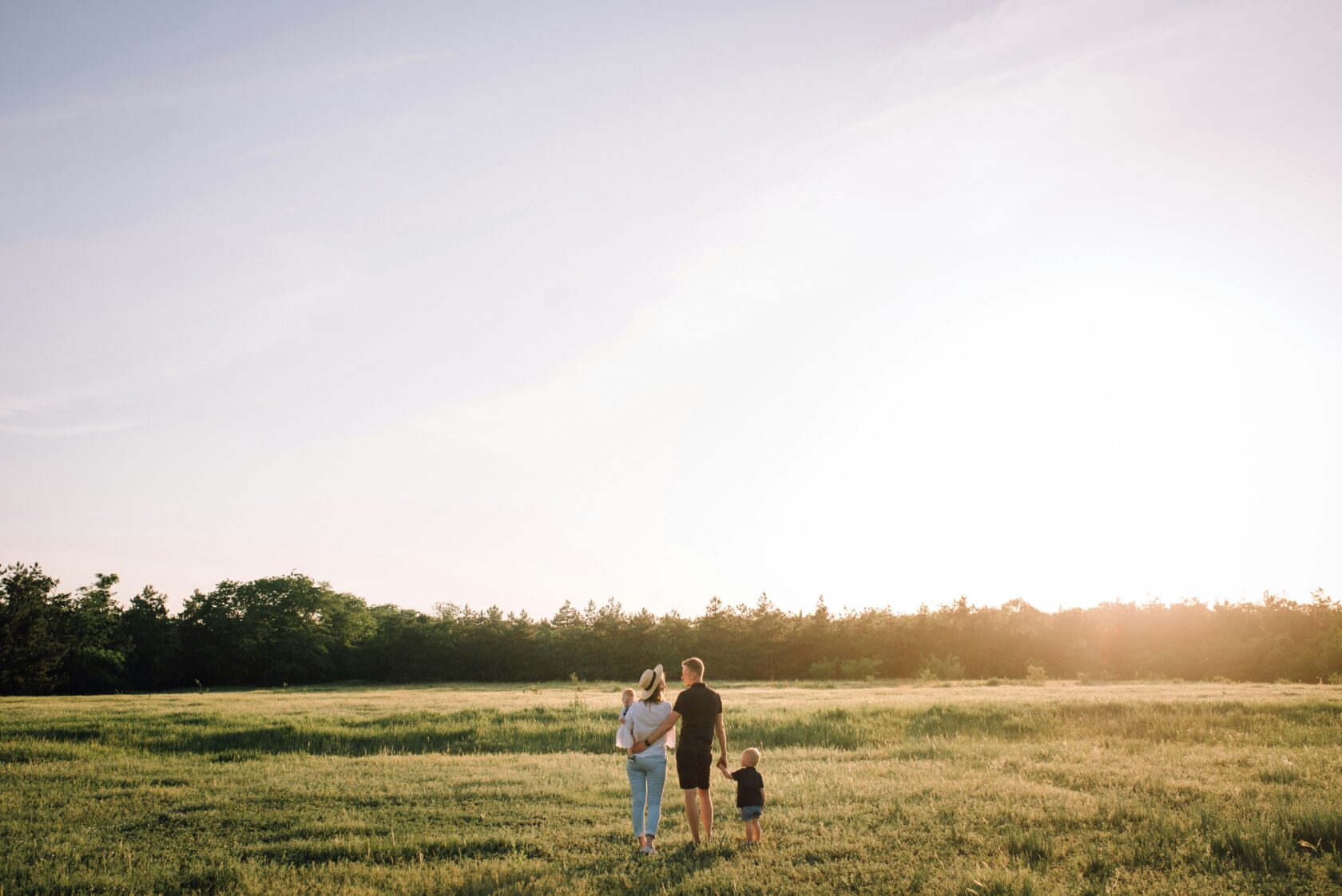 A family of four walking hand in hand through a sunlit field during sunset, with trees in the background.