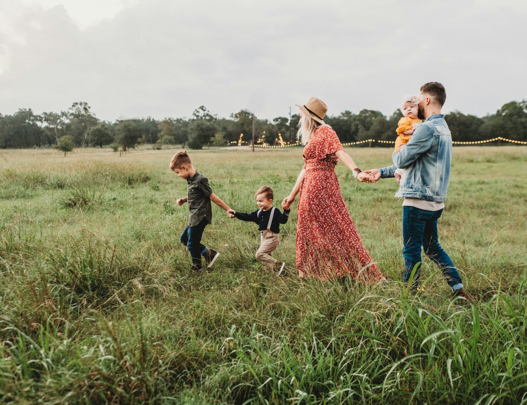 A family of five walking hand in hand through a grassy field, with two boys leading the way and a baby girl being carried by their father. The mother wears a red floral dress and a hat, while the background features trees and soft lighting.