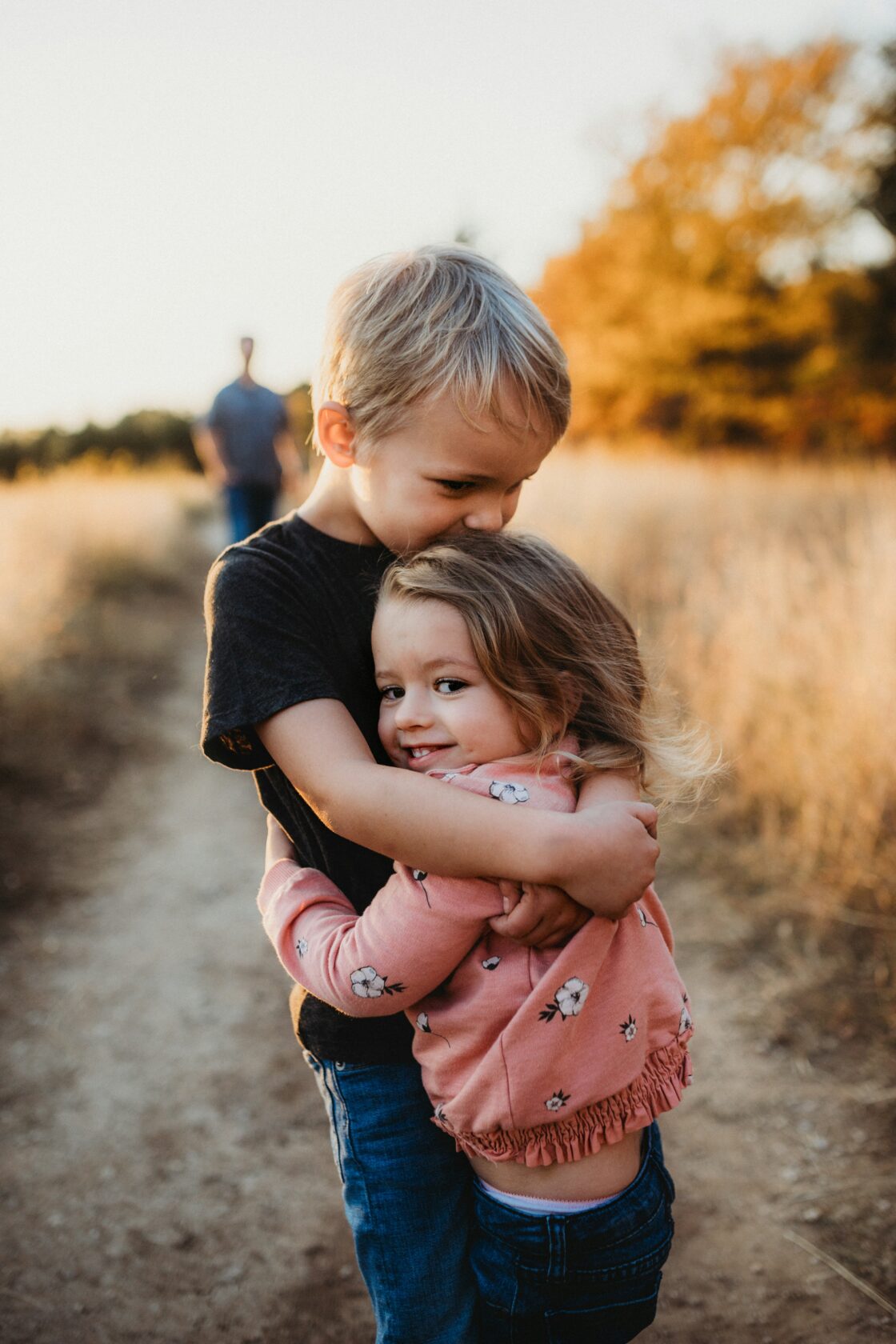 A young boy hugs his little sister from behind with a joyful expression, set against a golden backdrop of a sunlit field.