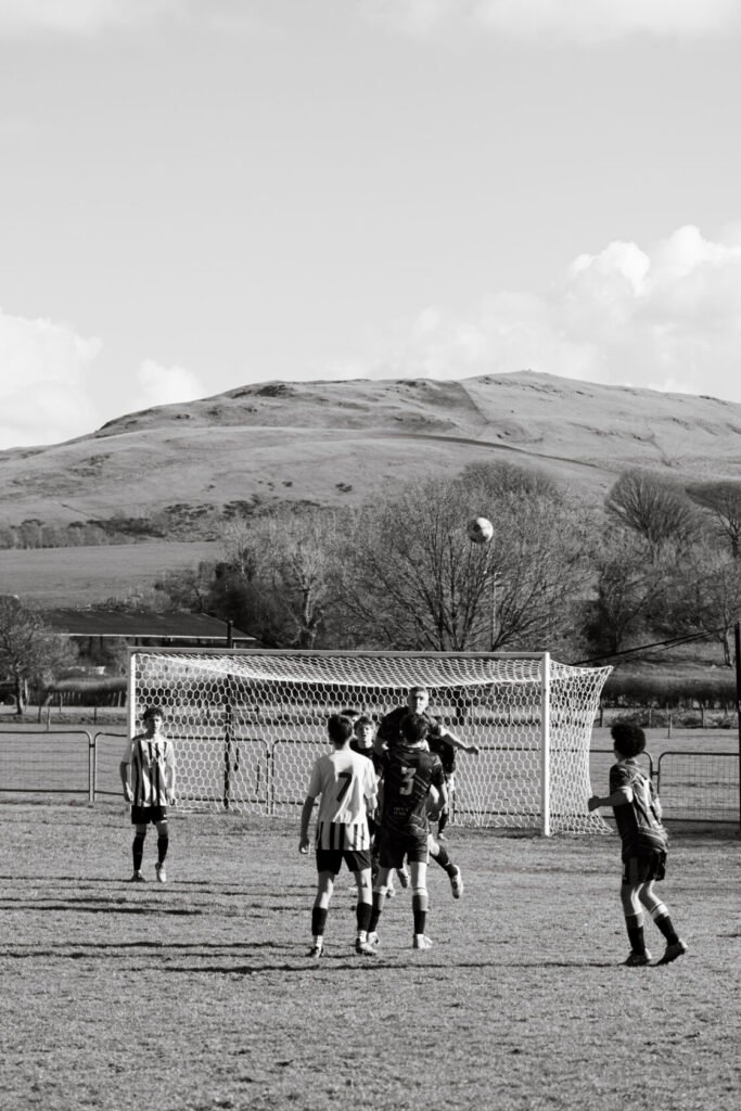 A black and white photograph of a youth football match, with players jumping to head the ball as it flies toward the goal, set against a backdrop of rolling hills and trees.