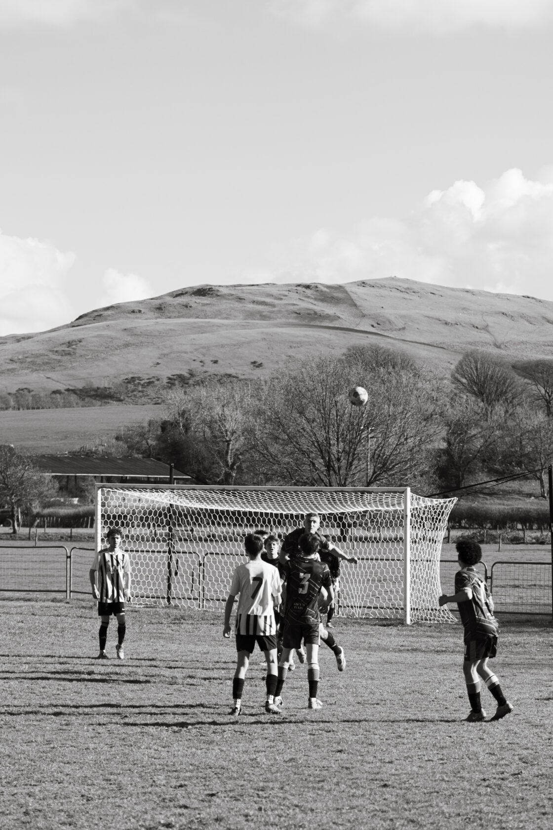 Youth Football Match with Scenic Hills A black and white photograph of a youth football match, with players jumping to head the ball as it flies toward the goal, set against a backdrop of rolling hills and trees.