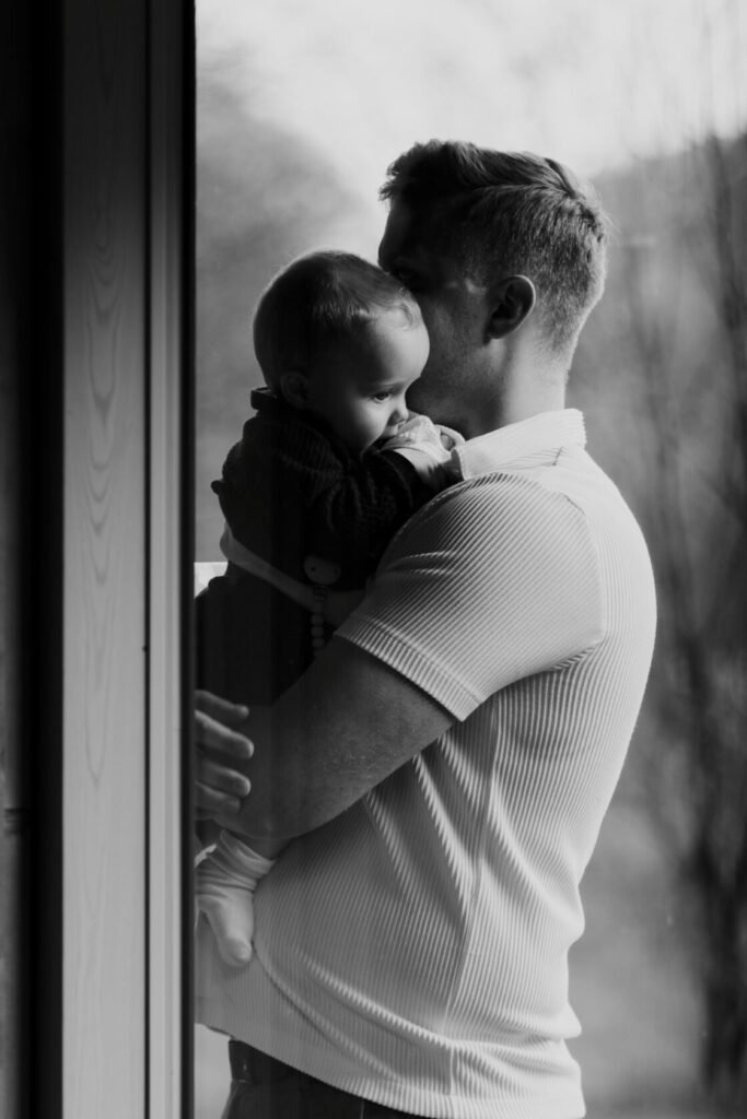 A black and white photograph of a father holding his baby close, nuzzling his forehead against the child's head, with a soft background suggesting a serene environment.
