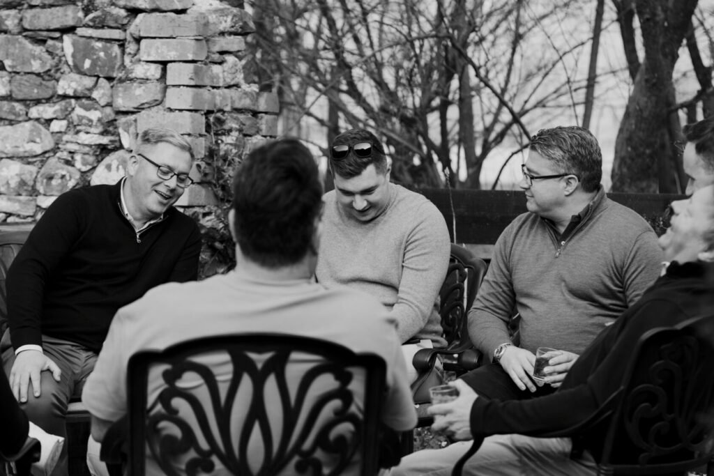 A group of men sitting together in an outdoor setting, smiling and interacting, with a rustic stone wall and trees in the background.