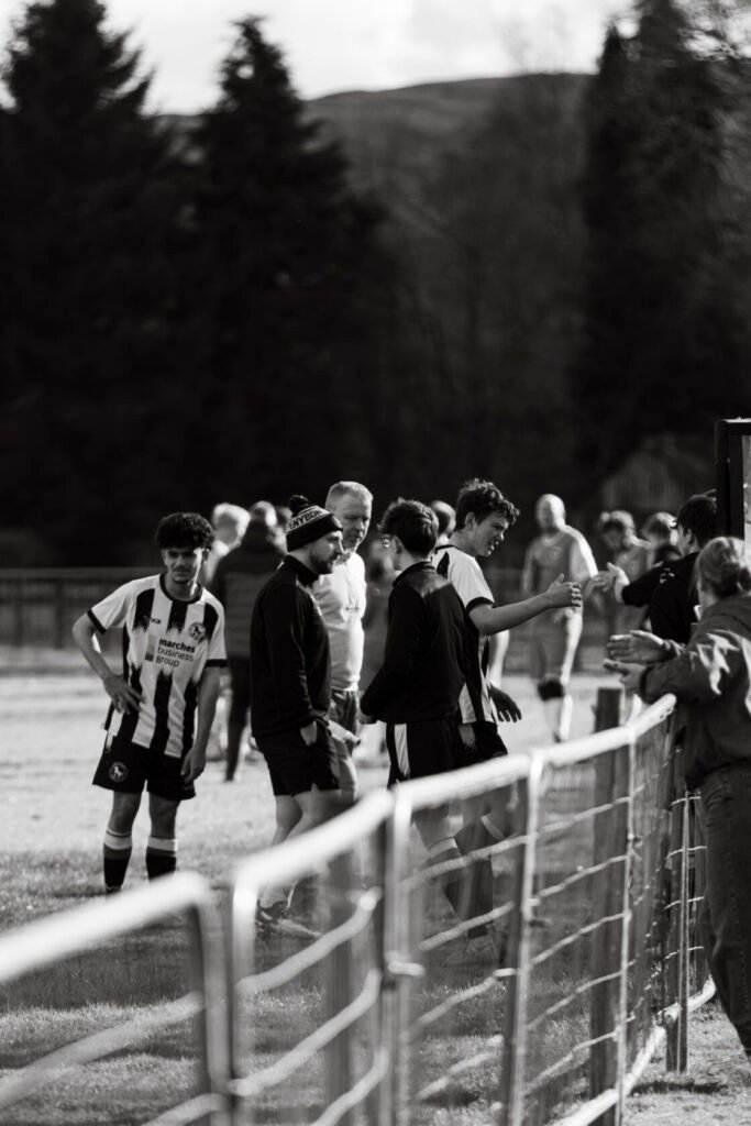 A group of young football players and coaches gathered near a pitch, dressed in team kits and casual clothing, engaged in conversation after a match.