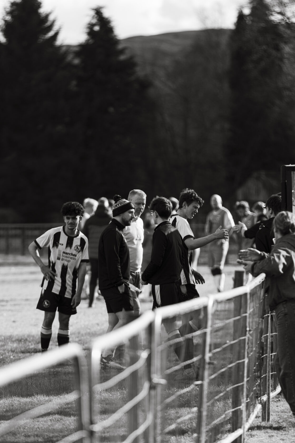 Youth Football Match Highlights A group of young football players and coaches gathered near a pitch, dressed in team kits and casual clothing, engaged in conversation after a match.