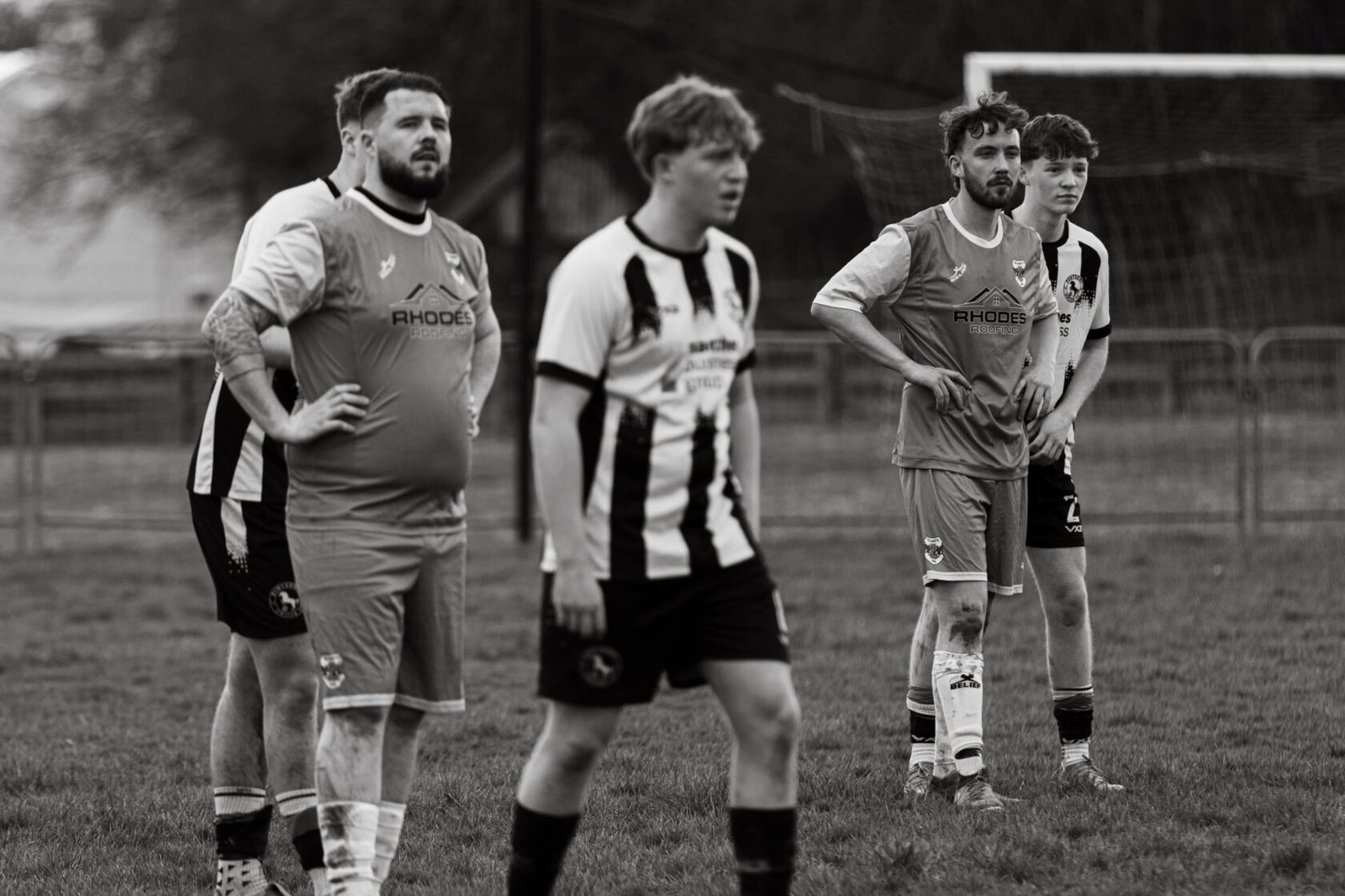 A group of football players in a black and white setting, two wearing grey jerseys and others in black and white striped uniforms, standing on the pitch with a focused expression.