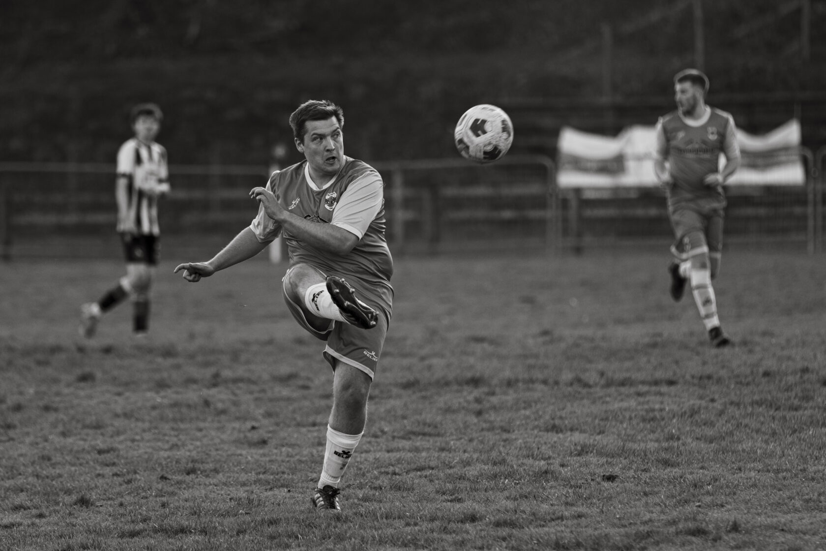Football Player Kicking Ball in Action A male football player wearing an orange and grey kit is executing a kick on the ball, while another player is in the background wearing a black and white striped kit.