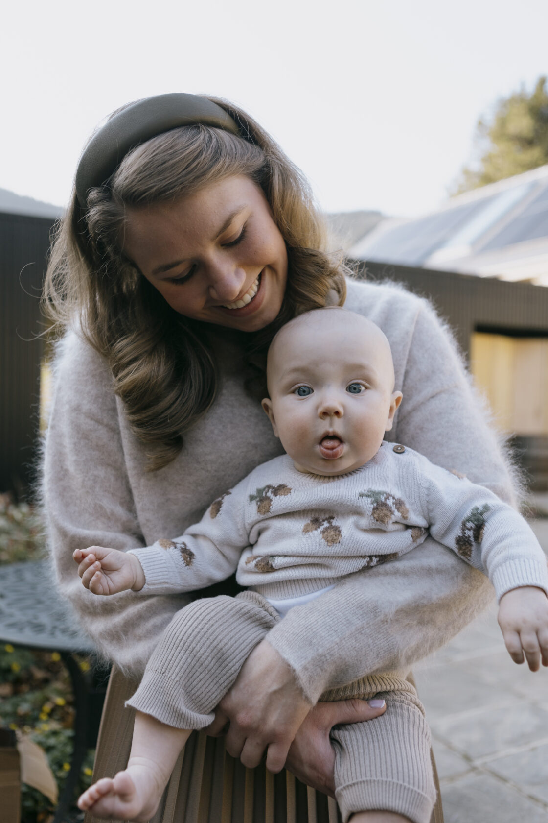 A smiling mother holds her baby outdoors, with the baby playfully sticking out their tongue. The mother has wavy hair and is wearing a cosy light sweater, while the baby is dressed in a patterned sweater and soft trousers.