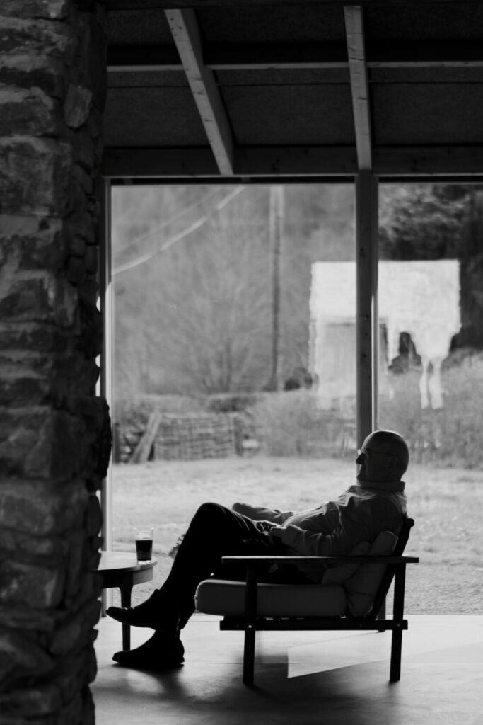 A man sitting in a modern chair in silhouette against a large window, with a rustic landscape visible outside. A small table beside him holds a glass, and the interior features stone walls.