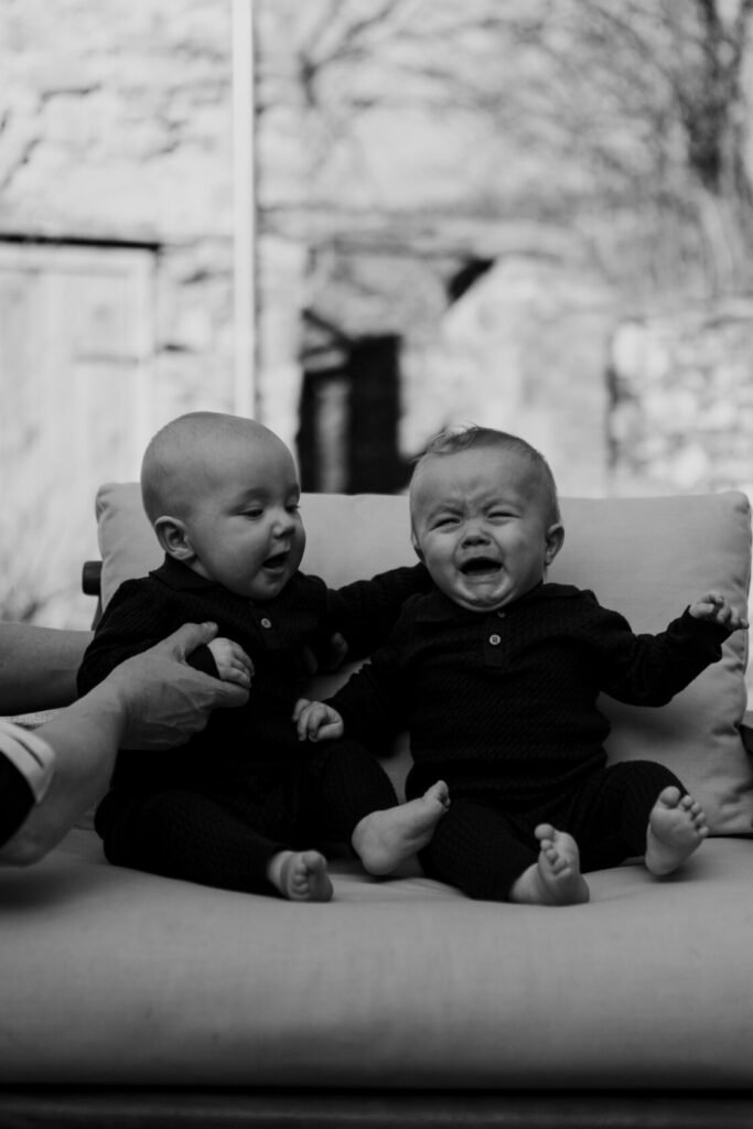 Two babies seated on a couch, one smiling and the other crying, with hands reaching towards them.