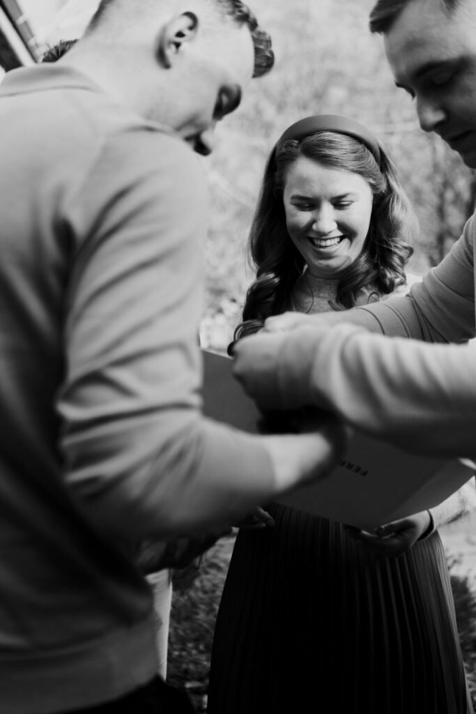 Three people, two men and a woman, gathered around a gift box. The woman is smiling while the men assist in unwrapping it. They are outdoors, with trees in the background and the scene is in black and white.