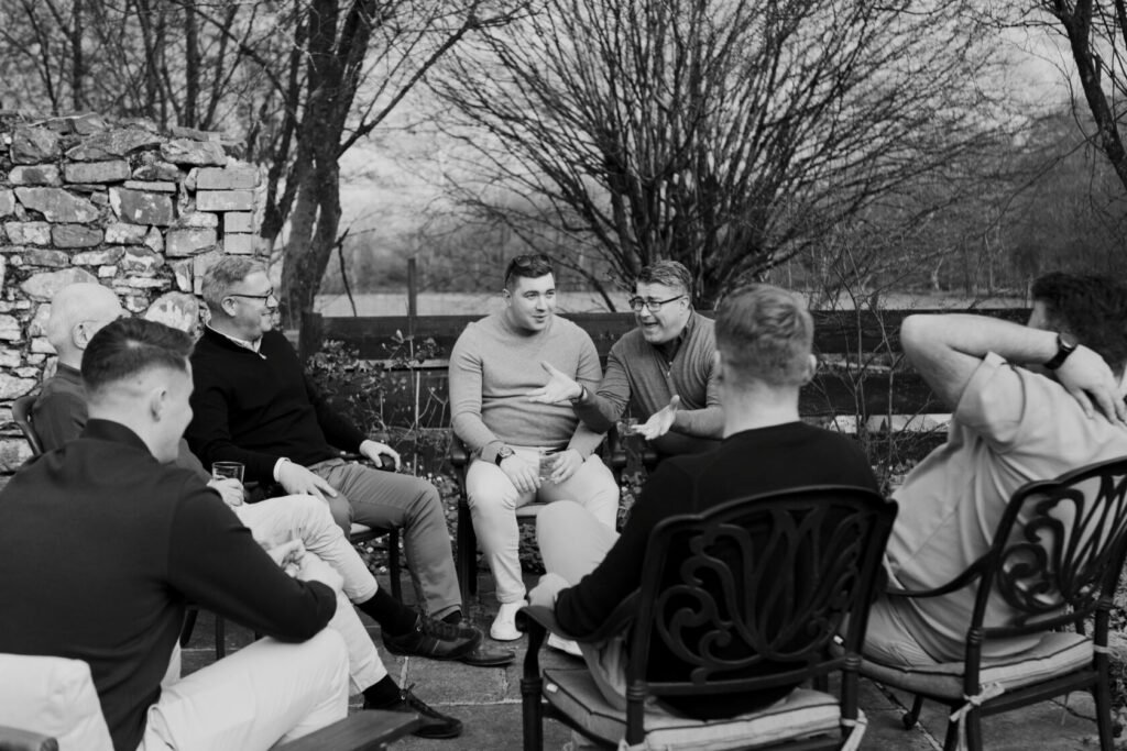 A group of men sitting in a circle on garden chairs, engaged in conversation. Some hold drinks, while others are animatedly listening. The background features a stone wall and bare trees.