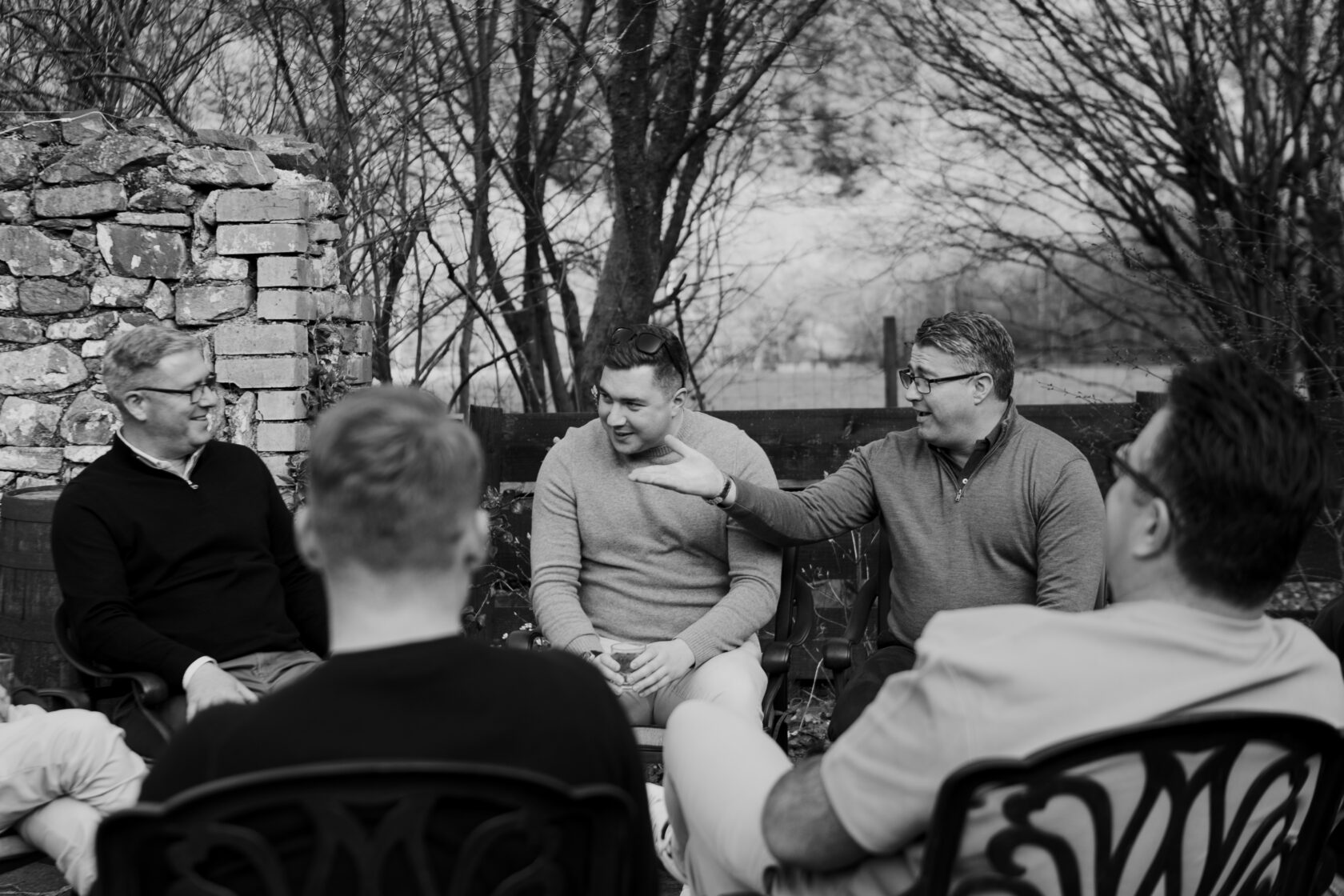 Group of men engaged in a relaxed discussion outdoors, with a stone wall and trees in the background. Two men are smiling and talking, while others listen attentively.