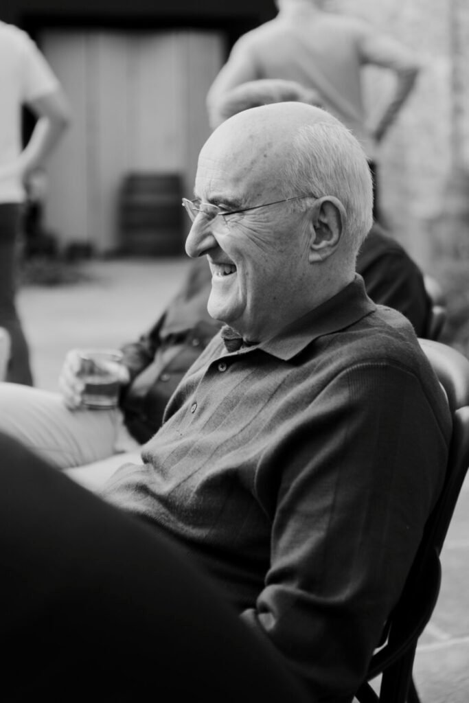 A smiling elderly man with a bald head and glasses, wearing a dark polo shirt, seated in a casual outdoor setting. In the background, blurred figures and barrels are visible.