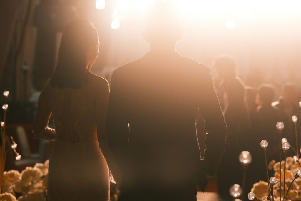 Silhouette of a couple holding hands, standing together at a wedding ceremony. The scene is illuminated by soft lights with flowers in the foreground.