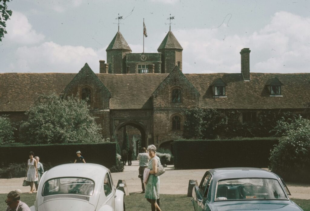 View of a traditional brick country house featuring turreted towers, surrounded by trees and hedges, with people walking in the foreground and two vintage cars parked in front.
