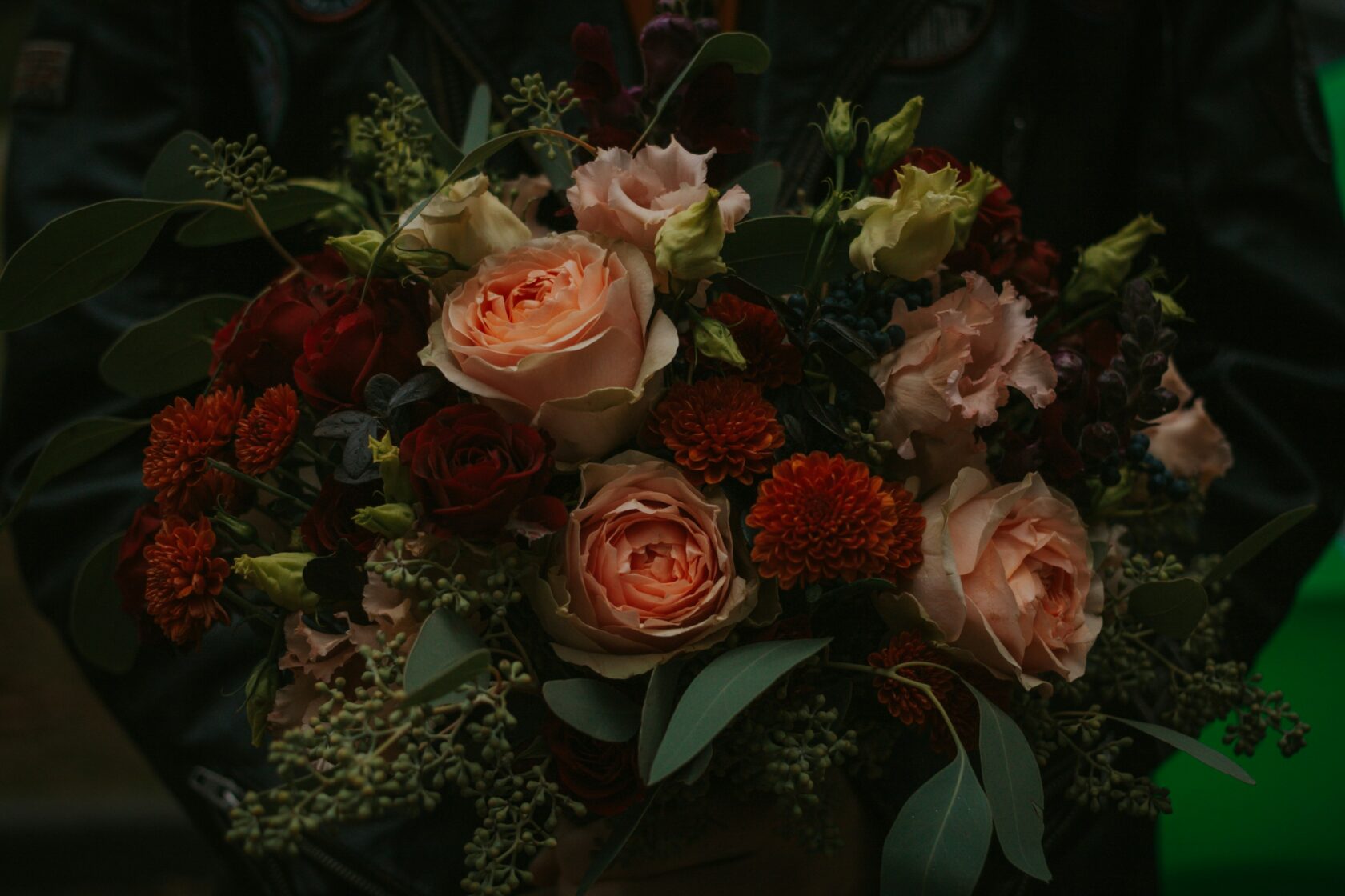 A close-up view of a lush bouquet featuring various flowers, including peach roses, deep red roses, marigolds, and greenery, held by a person wearing a black leather jacket.