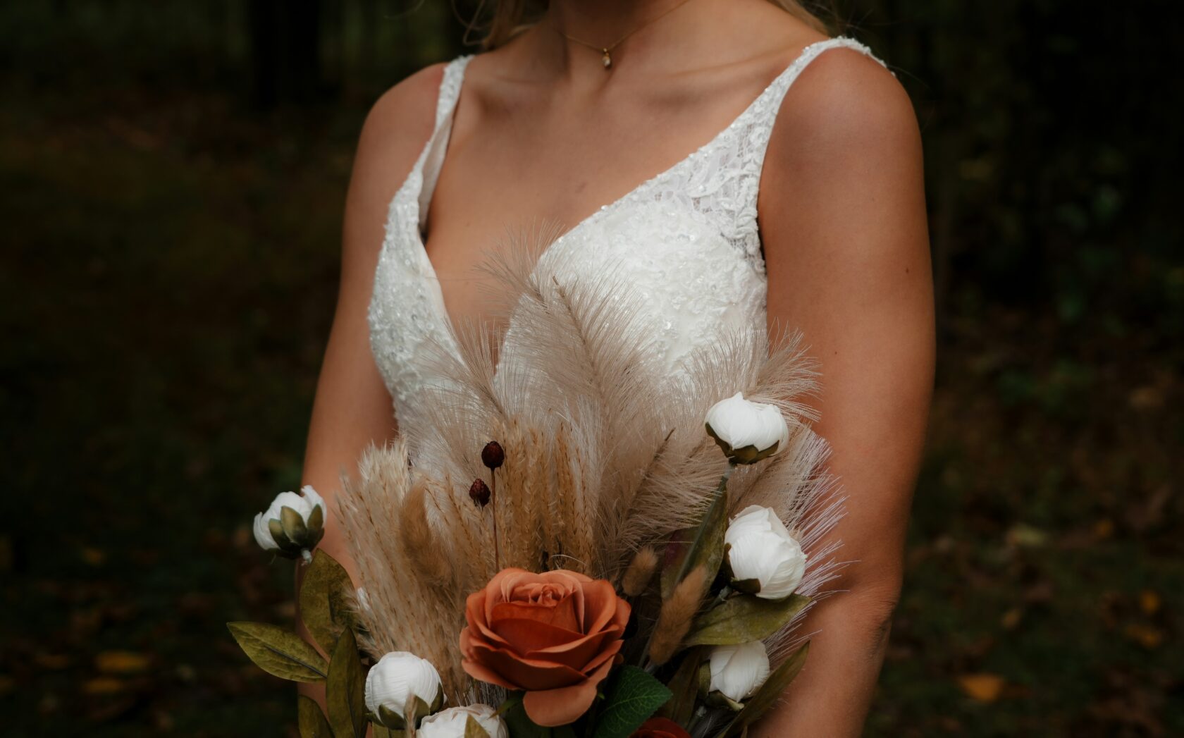 Close-up of a bride holding a bouquet featuring dried flowers and roses, wearing a lace wedding dress with spaghetti straps.