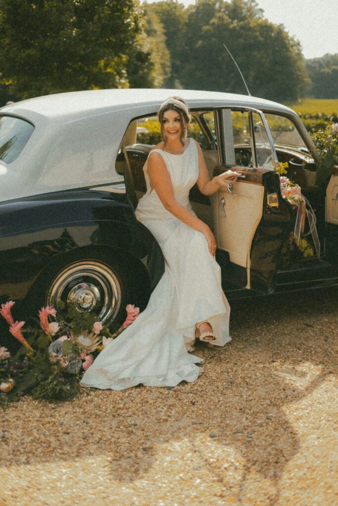 A smiling bride in a sleek white gown sitting on the step of a classic car, surrounded by floral arrangements and a scenic backdrop.