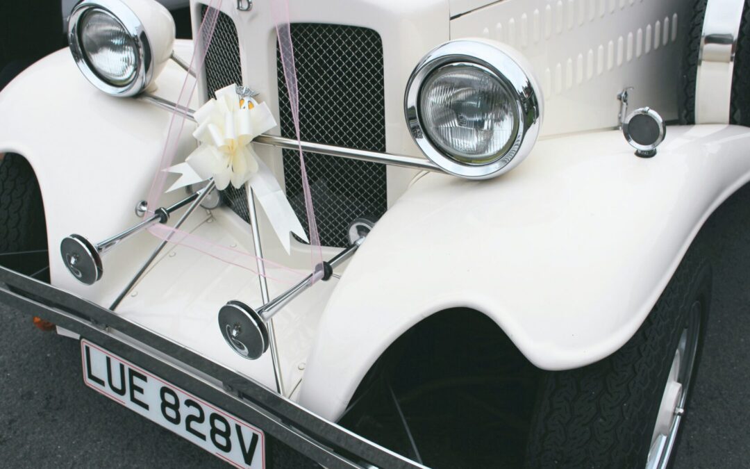 Close-up of a vintage white car's front with chrome details, featuring a decorative white bow and pink ribbons