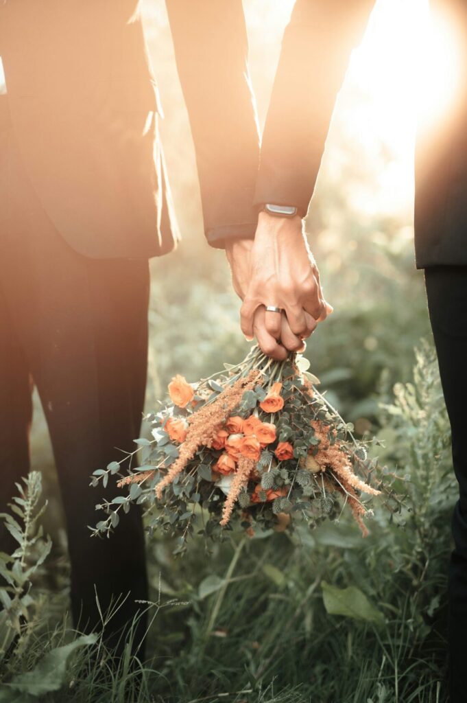 Two individuals in formal attire holding hands, with one person holding a bouquet of orange roses and greenery, set in a sunlit grassy field.