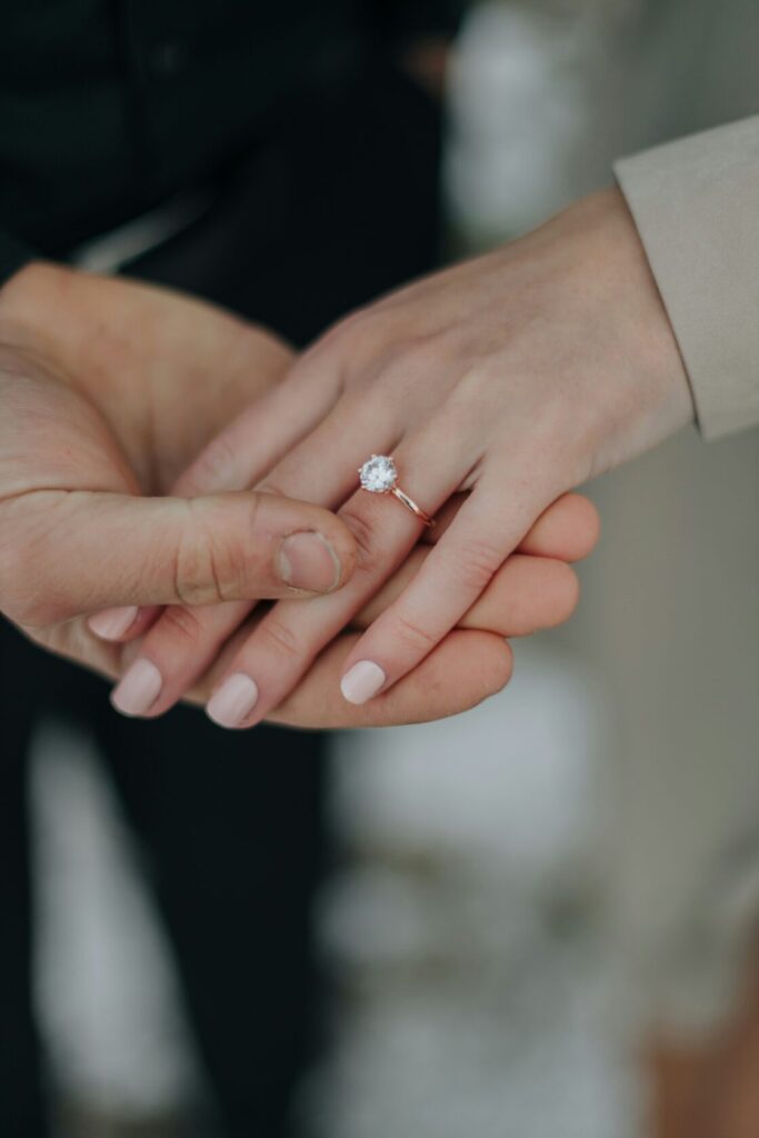 Close-up of two hands holding each other, featuring a diamond engagement ring on a woman's hand with light pink nail polish.