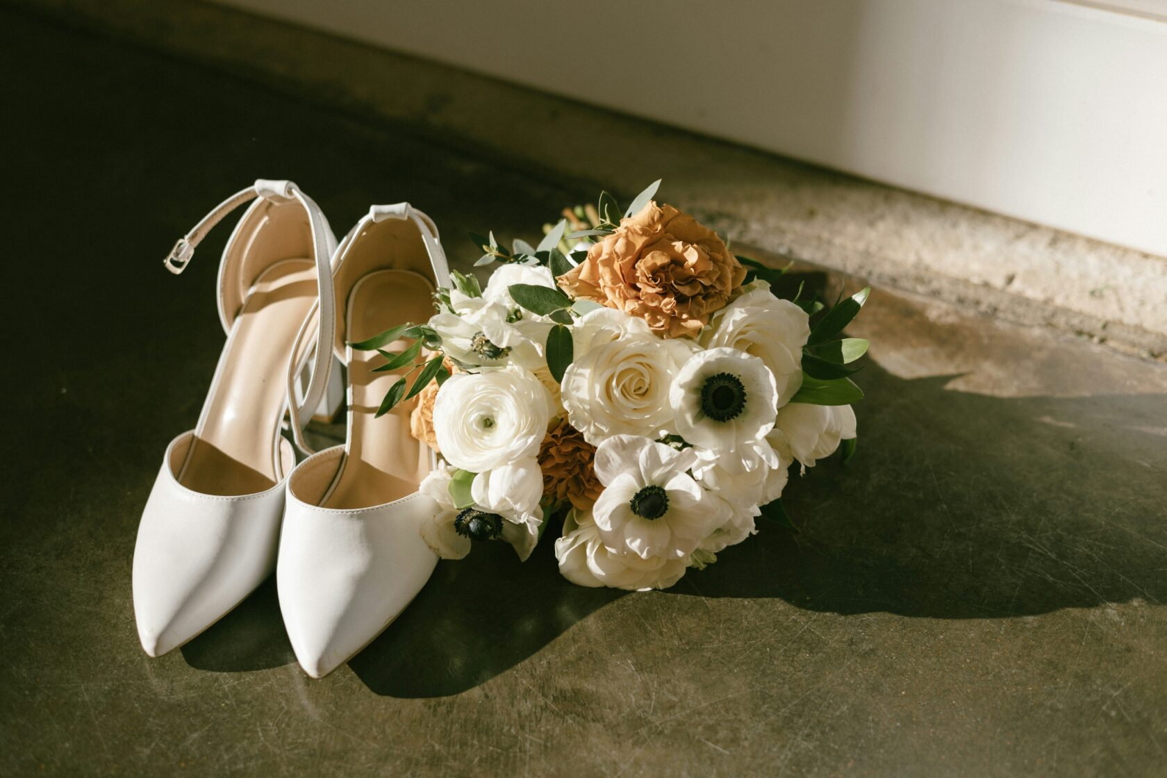 A pair of white pointed-toe shoes next to a bridal bouquet featuring white and peach flowers with greenery.