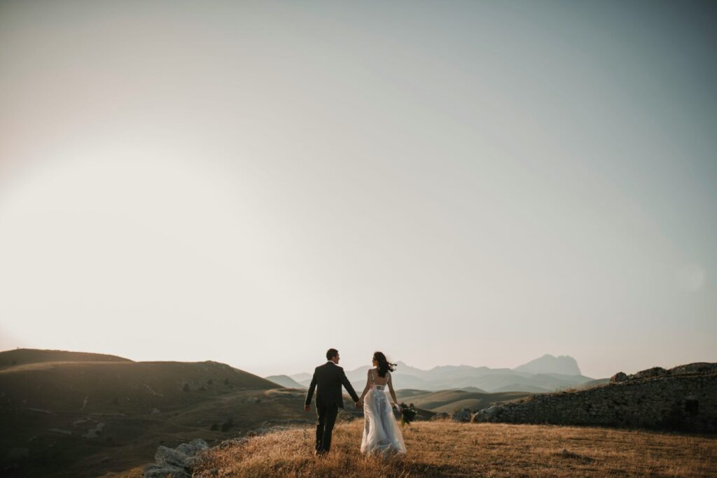 A newlywed couple walking hand in hand on a grassy hillside, with mountains in the background and a soft sunset glow.
