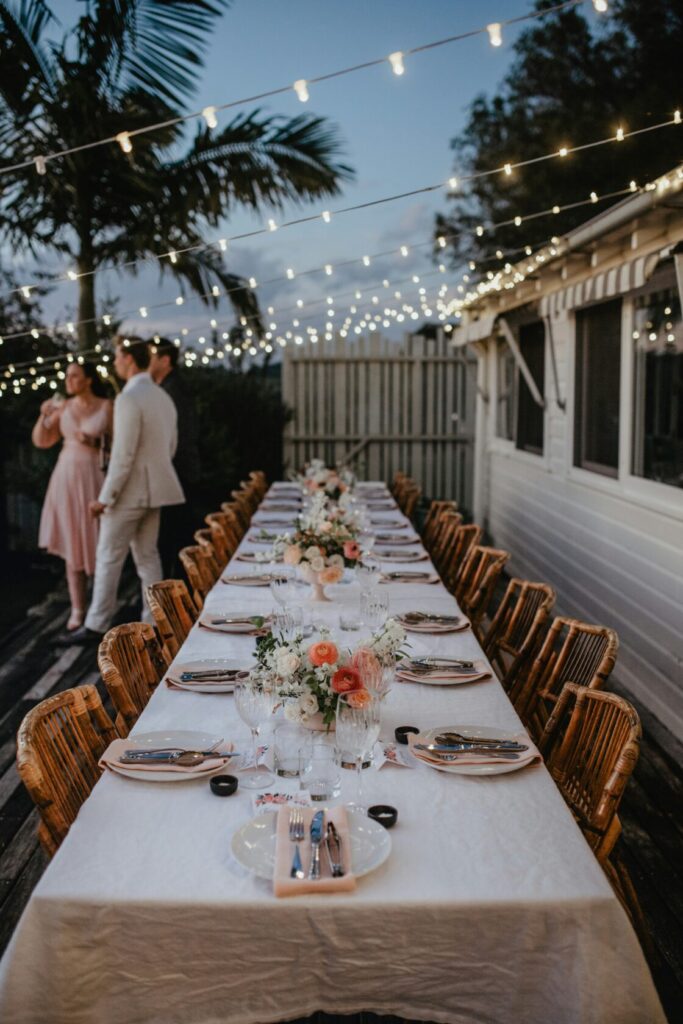Long white table set for an outdoor dinner with floral arrangements, glassware, and string lights overhead in a garden setting.