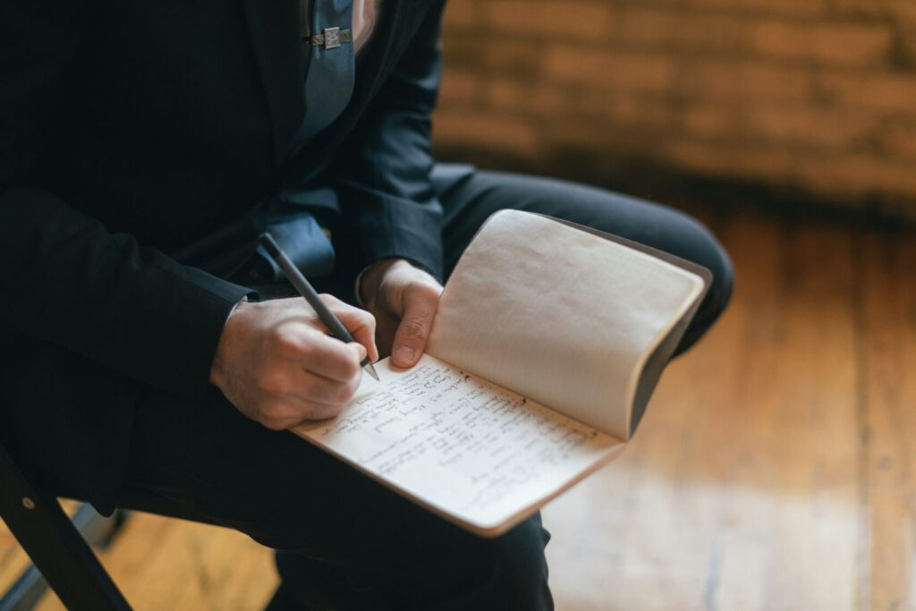 A man in a suit writing notes in a lined notebook while seated on a wooden chair, with a focus on his hands and the open notebook.