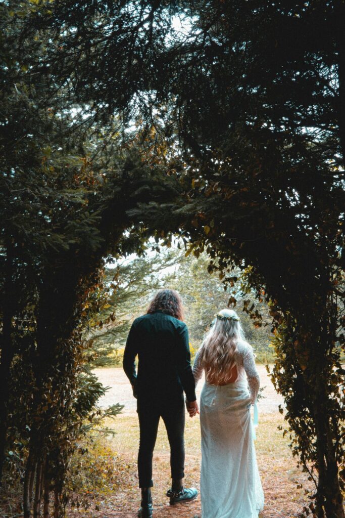 A couple holding hands while walking through an arch of trees. The woman wears a long white wedding dress with lace details and has long, flowing hair adorned with flowers. The man is dressed in black, with long curly hair.